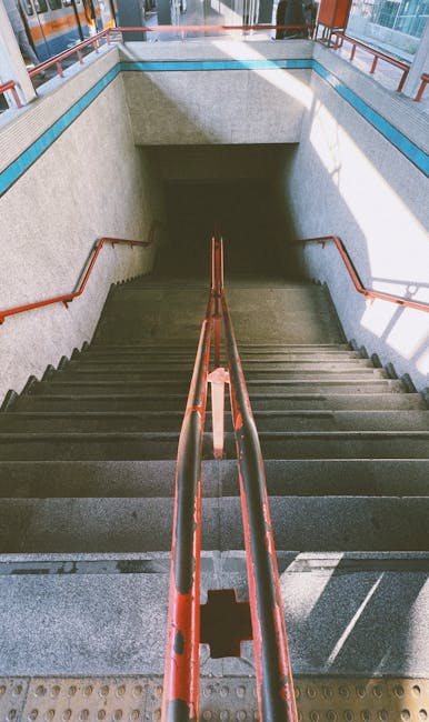 A concrete staircase with metal handrails painted in a reddish-brown color leading down to an underground level, with a blue-tiled wall at the top and a ceiling above the stairwell. The stairs are evenly spaced and illuminated by natural light from an opening at the top. In the context of house removals and furniture transport, such staircases can present access challenges during a home relocation, particularly for large furniture items. Lambeth Movers' services may include solutions for stair navigation, ensuring the safe and efficient loading or unloading of items through narrow or difficult staircases in properties like this. The environment appears clean and professional, emphasizing careful handling during the moving process.