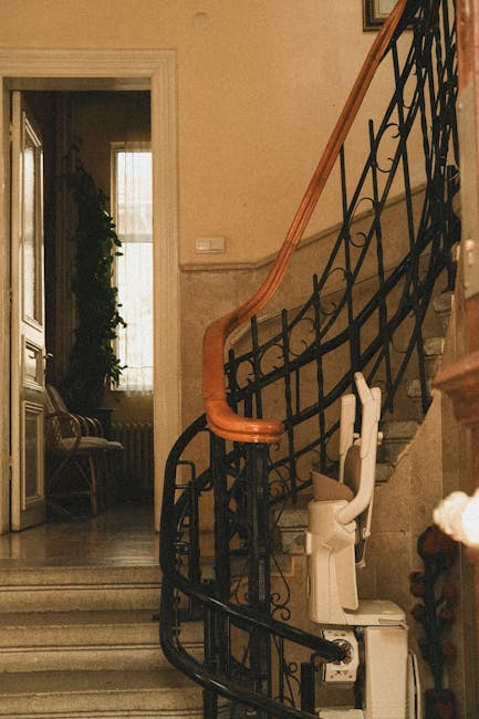 A narrow, indoor staircase in a residential property featuring ornate black wrought iron railing with a wooden handrail. The marble steps have a polished surface, and a stairlift with a beige seat and footrest is positioned along the side, indicating accessibility adaptations. Natural light filters in through a large window with sheer curtains in the background, illuminating the interior. To the left, part of a sitting area with a upholstered bench and decorative items is visible through an open doorway. The scene captures the environment for home relocation or furniture transport services, emphasizing the staircase's accessibility considerations. Lambeth Movers handles removals involving complex access issues, including staircase and staircase lift solutions, supporting efficient and safe moving logistics in SE11. The environment appears clean and well-maintained, with a focus on precise description of the staircase and surrounding interior.