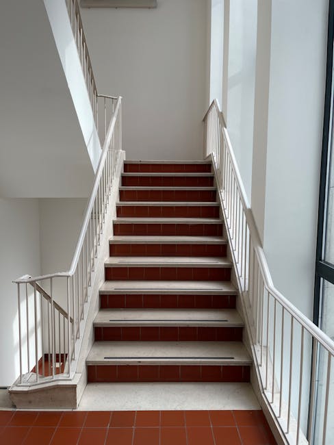 A concrete staircase with metal handrails painted in a reddish-brown color leading down to an underground level, with a blue-tiled wall at the top and a ceiling above the stairwell. The stairs are evenly spaced and illuminated by natural light from an opening at the top. In the context of house removals and furniture transport, such staircases can present access challenges during a home relocation, particularly for large furniture items. Lambeth Movers' services may include solutions for stair navigation, ensuring the safe and efficient loading or unloading of items through narrow or difficult staircases in properties like this. The environment appears clean and professional, emphasizing careful handling during the moving process.