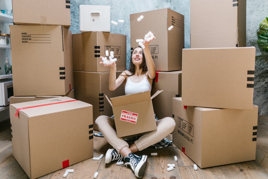 A young woman sitting cross-legged on a wooden floor inside a home, surrounded by numerous unpacked cardboard boxes of various sizes, some sealed with red tape and others open, with packing foam peanuts scattered around her. She is smiling and tossing foam packing peanuts into the air, with a partially open box nested on her lap. Behind her, a light blue textured wall and a plant on the right side are visible, indicating a room prepared for house moving or furniture transport. The scene captures the unpacking stage of home relocation, with some boxes labeled for contents and room designation, and reflects the logistical process involved in packing and moving items during a house move, such as a Victorian terrace in Brixton. The environment is well-lit, emphasizing the focus on packing materials, cardboard boxes, and the interior setting facilitated by Lambeth Movers during a furniture transport or house removals service.