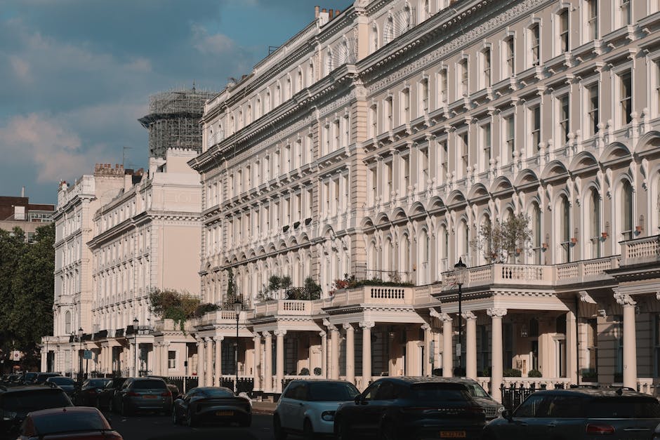 A row of Victorian-style terraced houses with white facades and decorative architectural details, including arched windows and columns, situated on a residential street in Brixton. The street features parked cars along the curb, with some vehicles partially obscuring the lower portions of the buildings. The scene is lit by daylight, with a partly cloudy sky overhead and a building in scaffolding visible in the background. In the context of house removals, furniture, boxes wrapped in plastic and cardboard, and moving equipment such as trolleys and straps might be seen in front of or near the entrance, indicating an active loading or packing process. Lambeth Movers, a professional removals company, often use such environments for furniture transport and home relocation services, supporting efficient packing and logistics during Victorian terrace moves in Brixton.