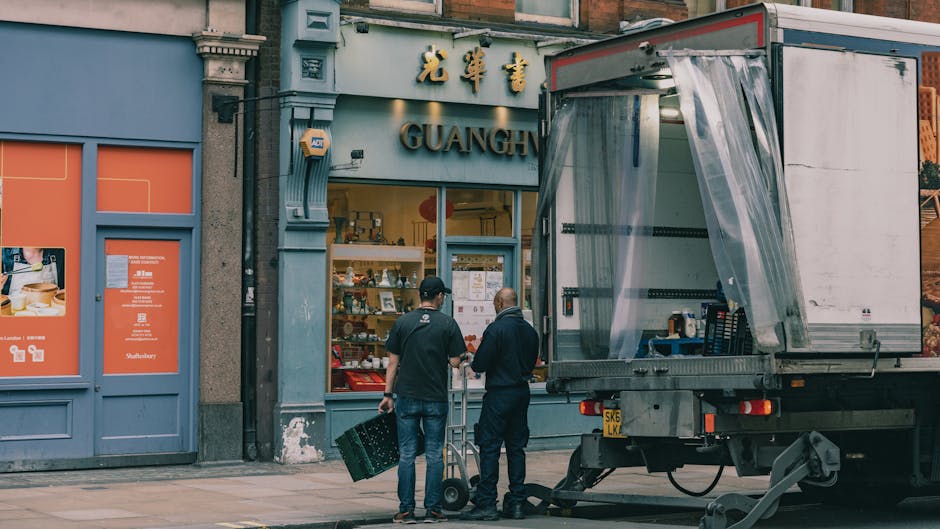 Two men standing outside a shop with Chinese signage, preparing for a home relocation or furniture transport, as a moving truck with an open side panel and transparent plastic curtain is parked nearby on the pavement. The man on the left is holding a black carrying case, while the man on the right is pushing a hand trolley. Inside the truck, partially visible, are packing materials such as cardboard boxes and wrapping items, with some furniture or appliances possibly being loaded or unloaded. The scene is set in an urban area with a storefront displaying various items through large glass windows, and the exterior of the building features traditional architectural details. Lighting indicates daytime, with natural light illuminating the scene, reflecting typical moving procedures involved in packing and furniture transport during a house removal coordinated by Lambeth Movers.