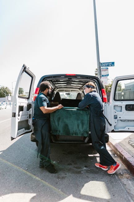 Two professional movers from Lambeth Movers are engaged in loading a large, dark green upholstered piece of furniture into the open cargo area of a white van parked on a street. One mover, wearing a navy jacket and black trousers, holds the furniture steady, while the other, dressed in a blue t-shirt and black pants, assists with guiding it into the vehicle. The van’s rear doors are wide open, revealing a spacious interior intended for transporting household items. The scene takes place outdoors during daytime, with bright natural light illuminating the setup, and the background features a street sign indicating Somerset Road, a nearby sidewalk, and a few parked cars. The movers are equipped with straps and blankets to safeguard furniture during home relocation, and the setting reflects the loading process typical of furniture transport services involved in house removals or moving logistics, emphasizing the professional handling of household items by Lambeth Movers.