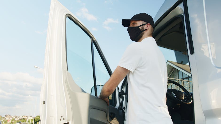 A male removal specialist from Lambeth Movers, wearing a black face mask, black cap, and white t-shirt, is standing inside the cab of a large white moving van with the door open. He appears to be preparing or inspecting the vehicle for home relocation or furniture transport. The interior of the cab shows a steering wheel and driver’s seat, while the background features a clear blue sky with some clouds and a residential area with houses and trees. The scene captures the loading process for a house removal service, emphasizing professional packing, moving logistics, and vehicle loading in an outdoor environment.