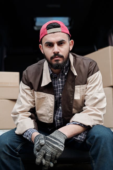 Two professional movers from Lambeth Movers are engaged in loading a large, dark green upholstered piece of furniture into the open cargo area of a white van parked on a street. One mover, wearing a navy jacket and black trousers, holds the furniture steady, while the other, dressed in a blue t-shirt and black pants, assists with guiding it into the vehicle. The van’s rear doors are wide open, revealing a spacious interior intended for transporting household items. The scene takes place outdoors during daytime, with bright natural light illuminating the setup, and the background features a street sign indicating Somerset Road, a nearby sidewalk, and a few parked cars. The movers are equipped with straps and blankets to safeguard furniture during home relocation, and the setting reflects the loading process typical of furniture transport services involved in house removals or moving logistics, emphasizing the professional handling of household items by Lambeth Movers.