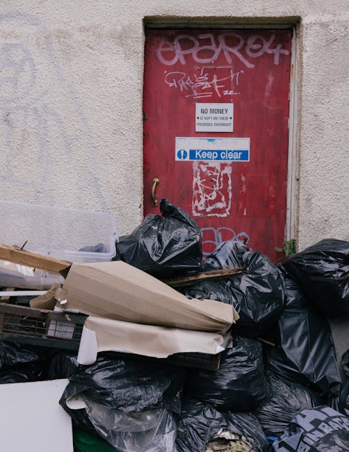 Outside a building with a weathered, beige wall and a red metal door covered in white graffiti tags and stickers, including a 'No Money' sign and a 'Keep Clear' notice, there is a pile of black plastic garbage bags filled with waste or unwanted items, situated on a pavement area next to the door. Some packaging materials, including crumpled cardboard and foam padding, are leaning against the bags. The scene suggests clearing or disposal activities, possibly linked to a home relocation or house clearance service by Lambeth Movers, with the presence of the bags and packaging indicating packing, loading, or waste removal during a move or property decluttering process.