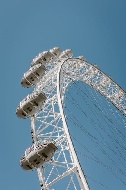 A large white Ferris wheel with multiple enclosed passenger cabins ascending in a vertical line, set against a clear blue sky. The structural framework of the wheel is visible, showing metal support beams and spokes that connect the central hub to the outer rim. The cabins are evenly spaced along the circumference of the wheel and are designed for passenger transport in an amusement park or fairground setting. The perspective of the photograph emphasizes the height and engineering details of the Ferris wheel, with no other objects or surroundings visible. This image captures the amusement ride in a static phase during daylight, relevant to topics such as transportation, entertainment installations, or large-scale leisure attractions, similar to the logistical considerations involved in home relocation or furniture transport for house removals or move planning.