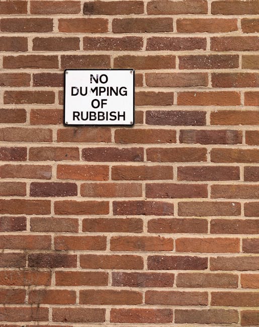 Outside a building with a weathered, beige wall and a red metal door covered in white graffiti tags and stickers, including a 'No Money' sign and a 'Keep Clear' notice, there is a pile of black plastic garbage bags filled with waste or unwanted items, situated on a pavement area next to the door. Some packaging materials, including crumpled cardboard and foam padding, are leaning against the bags. The scene suggests clearing or disposal activities, possibly linked to a home relocation or house clearance service by Lambeth Movers, with the presence of the bags and packaging indicating packing, loading, or waste removal during a move or property decluttering process.