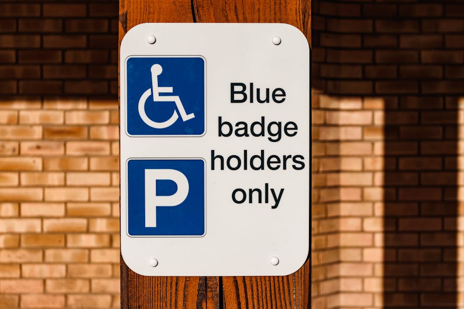 A rectangular outdoor sign mounted on a wooden post in front of a brick wall, indicating parking restrictions for disabled badge holders. The sign features a blue background with a white wheelchair symbol at the top, followed by the text 'Blue badge holders only' in black. Below, there is a large white letter 'P' on a blue background, denoting parking. The sign is positioned at a height suitable for visibility, and the surrounding environment suggests an area adjacent to a residential or commercial property, relevant for home relocation and furniture transport services. The lighting appears natural, casting subtle shadows on the sign and wall, with the sign serving as guidance for parking during a moving process or household removal coordinated by Lambeth Movers according to local parking rules for removals.