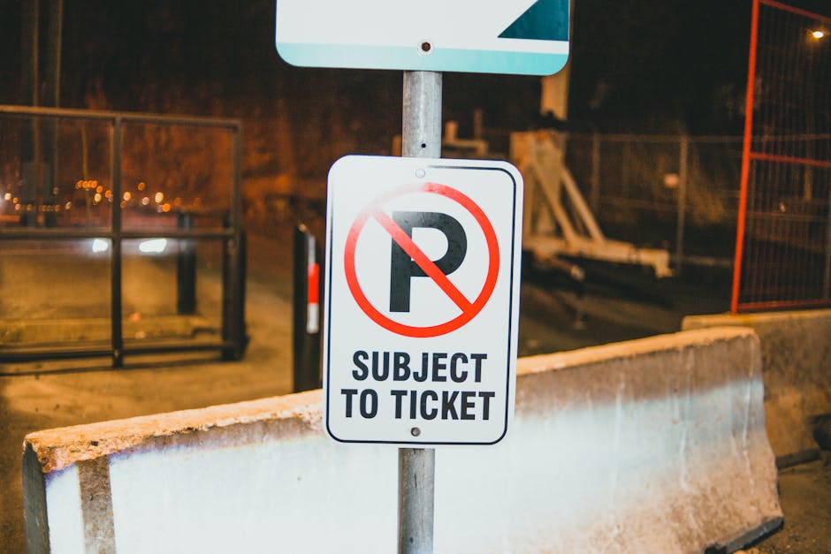 A street scene at night features a white parking restriction sign mounted on a metal pole, indicating no parking with a red circle and slash through a black 'P'. Below the symbol, the sign states 'SUBJECT TO TICKET' in bold black letters. The sign is positioned behind a concrete barrier on the pavement, with additional parking area markings or barriers visible in the background. The surroundings are dimly lit, with warm yellow street lighting illuminating the scene. This image captures the urban environment where house removals or furniture transportation may require adherence to parking regulations, which Lambeth Movers can help navigate during house relocation or packing and moving services.
