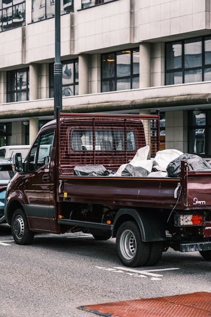 A small dark red flatbed van is parked on a city street in front of a modern office building with large glass windows and a concrete facade. The van's cargo area is filled with various household items including wrapped furniture pieces covered with white and black protective blankets, cardboard boxes, and plastic wrapping materials. A metal grid partition is installed behind the cab to secure the load. The items appear to be in the process of being loaded or unloaded, possibly as part of a home relocation or furniture transport service. The surrounding environment includes other parked vehicles, a streetlamp, and a paved sidewalk, indicating an urban setting suitable for house removals or same-day moving services. The van is positioned close to the curb, with the loading process visible, reflecting typical logistics involved in furniture moving or packing and moving operations conducted by Lambeth Movers.