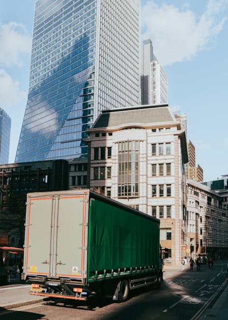 A large green and beige delivery truck with closed rear doors is parked on a city street, adjacent to modern high-rise office buildings with glass facades reflecting the blue sky and surrounding architecture. The truck is positioned on the road, next to a sidewalk where a few pedestrians and cyclists are visible, indicating an urban environment suitable for home relocation or furniture transport. The truck appears ready for loading or unloading, which is part of an ongoing house removal process. The scene is well-lit with natural daylight, emphasizing the urban setting typical of a professional moving service such as Lambeth Movers, who specialise in same-day removals to minimise delays and extra fees during house moves or furniture transport in Lambeth.