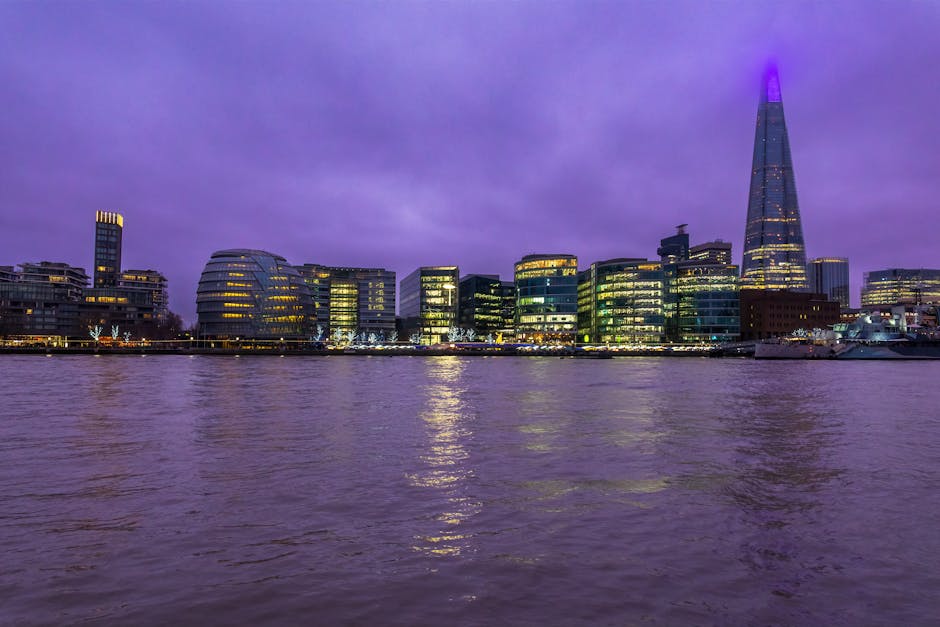 A nighttime cityscape of London’s South Bank showing modern office buildings and the distinctive Shard skyscraper illuminated in purple light. The buildings are situated along the riverbank, with their glass facades reflecting in the calm water below. The sky overhead is filled with purple-tinged clouds, creating a dramatic backdrop. In the foreground, the river’s surface appears gently rippled, with reflections of the illuminated windows and city lights. This scene captures the vibrant urban environment characteristic of London’s riverside, relevant to home relocation and furniture transport services such as those provided by Lambeth Movers, especially during the loading process of a house removal along the South Bank.