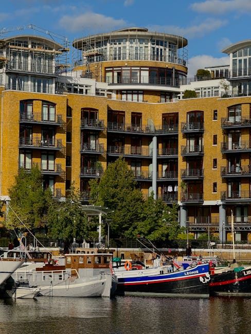 This image shows a multi-storey modern residential building with a curved architectural design, featuring large glass windows and multiple balconies, situated along a waterway in Lambeth. In front of the building, there are several boats docked at the riverbank, including a boat with the name 'LEONI' painted on its side, and another partially visible vessel. The foreground includes a wooden walkway or quay with some railings, and a few trees with green foliage positioned at the base of the building, contributing to a natural urban setting. The scene is captured during daylight hours with a partly cloudy sky, highlighting the vibrant urban lifestyle typical of South Bank, close to the Thames. Lambeth Movers, specialists in house and furniture removals, often operate in such environments, assisting with home relocation and furniture transport across SE1 flats and nearby areas, where loading and packing often involve careful navigation around boats and outdoor furniture.